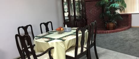 A dining area with a round table covered by a tablecloth, surrounded by wooden chairs. A glass bowl with fruits is on the table, and a mirror hangs on the wall.