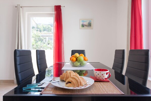 A dining table with chairs, a plate with croissants, a bowl of fruit, and a polka-dotted cup.