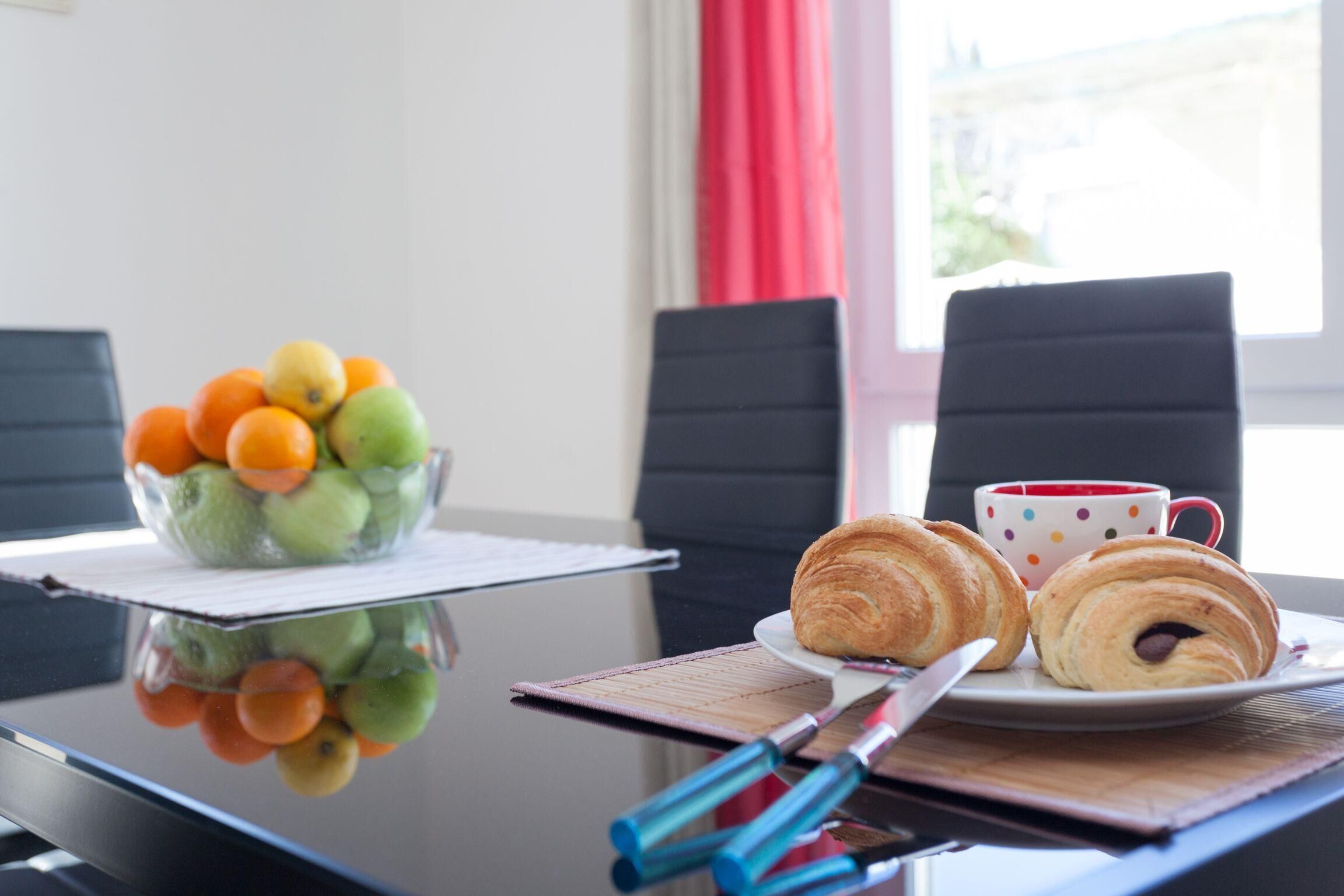 A table with a fruit bowl, croissants, a cup, and a knife.