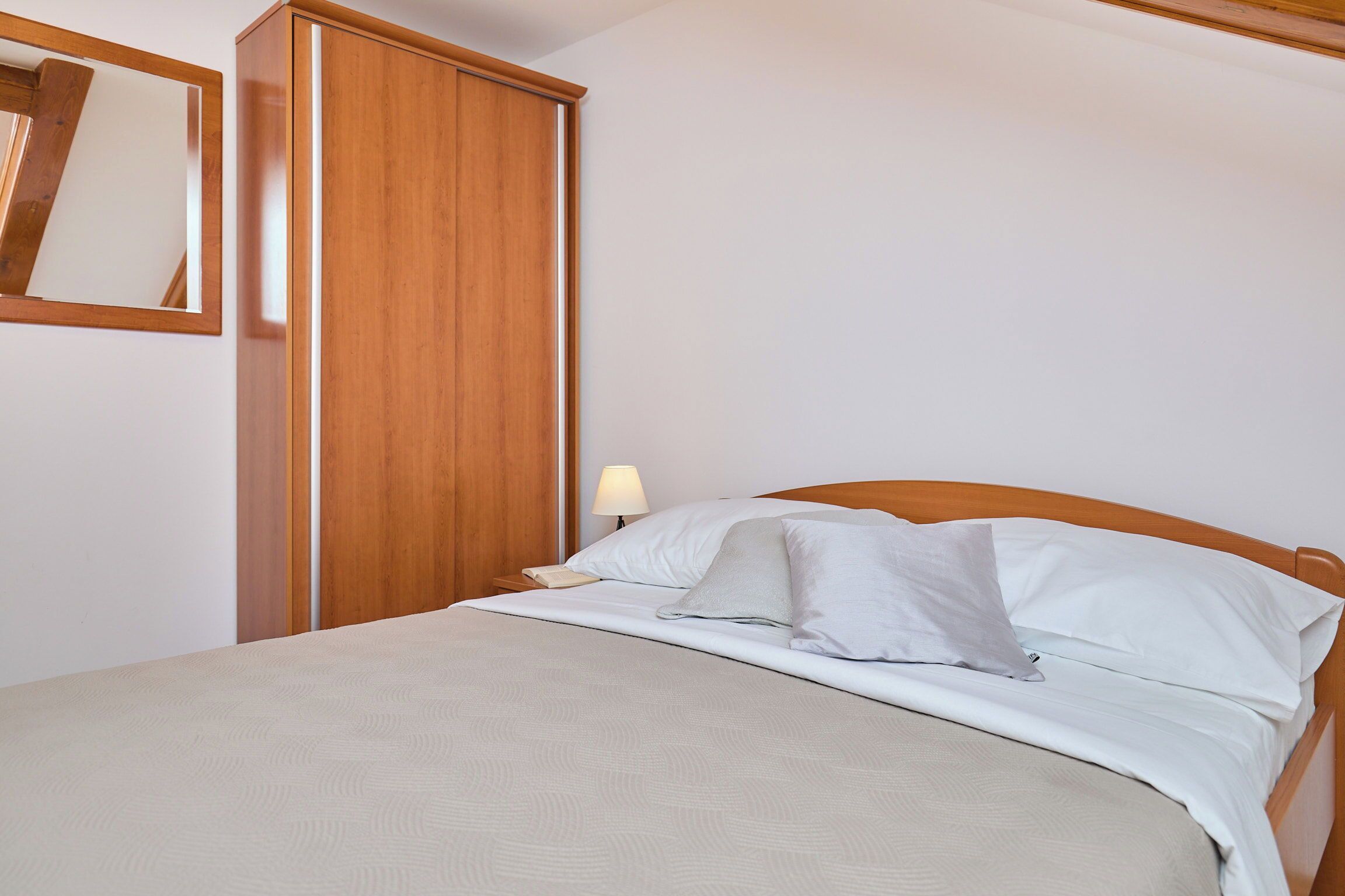 Una cama bien hecha con sábanas blancas y una manta color beige, un armario de madera, una lámpara de noche y un espejo en la pared.