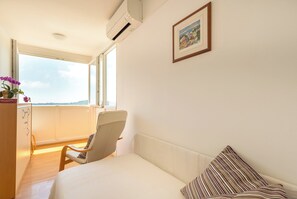 A bedroom with a bed, a chair, a desk, a window with a view, and an air conditioner.