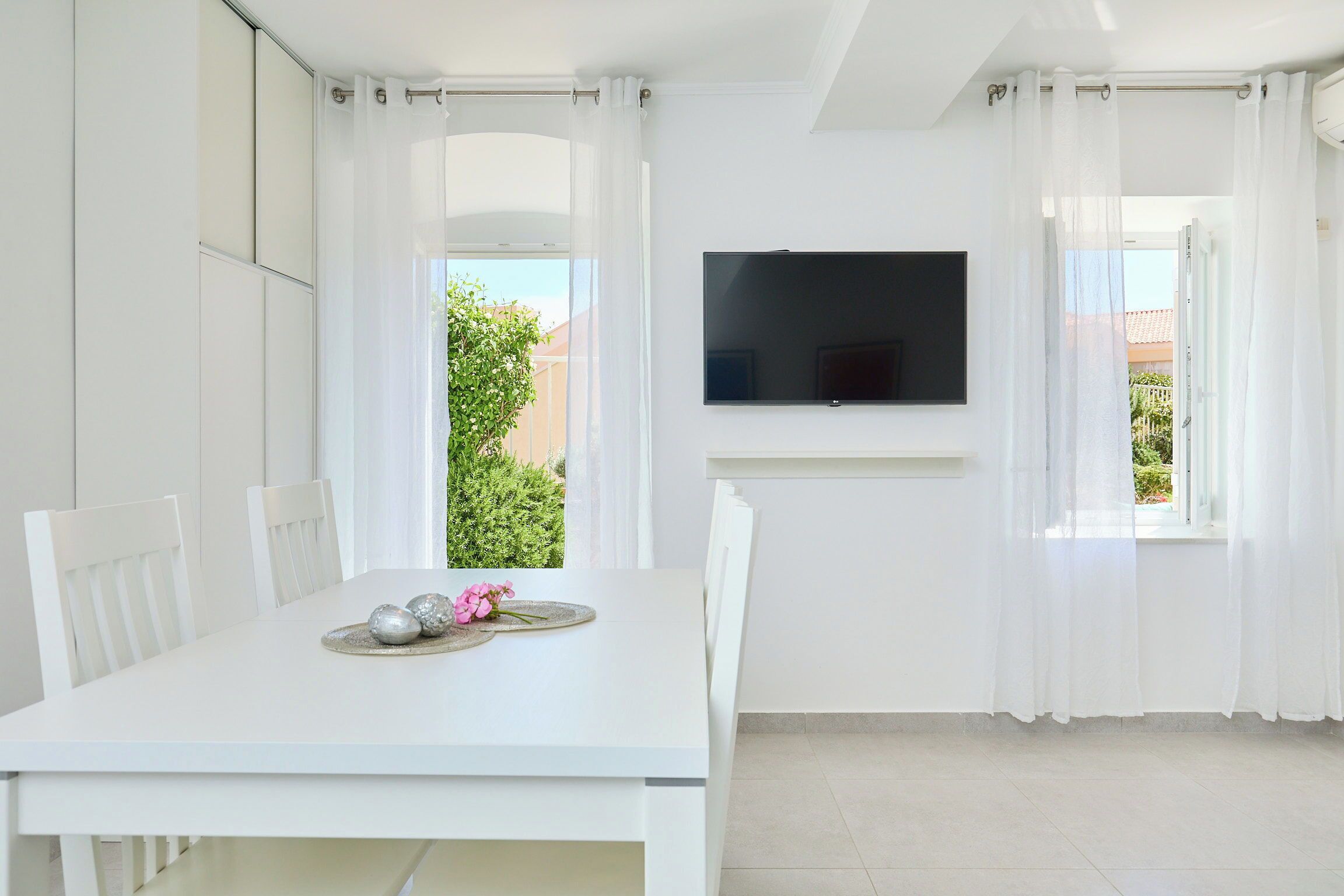 A modern, white room with a dining table, chairs, a flat-screen TV, and large windows with sheer curtains.