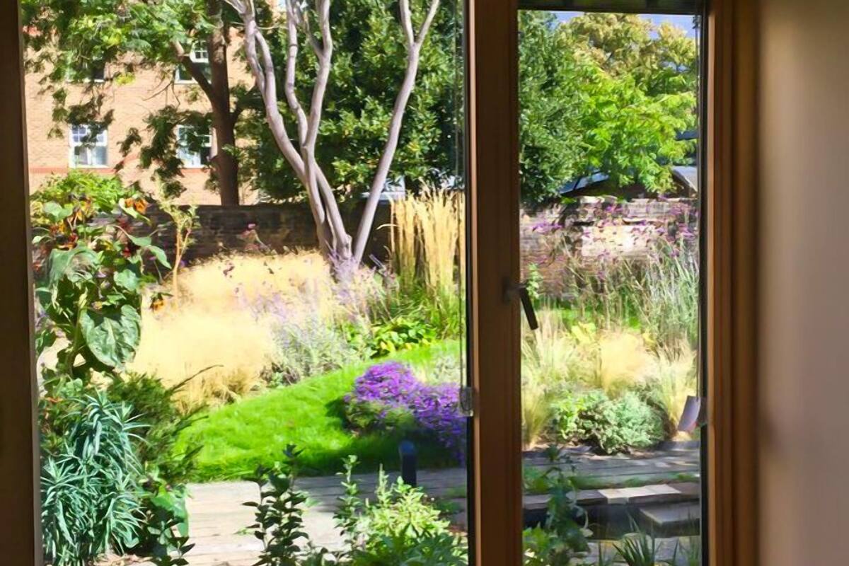 Ein Zimmer mit einem großen Fenster, das auf einen Garten mit verschiedenen Pflanzen und Bäumen blickt.