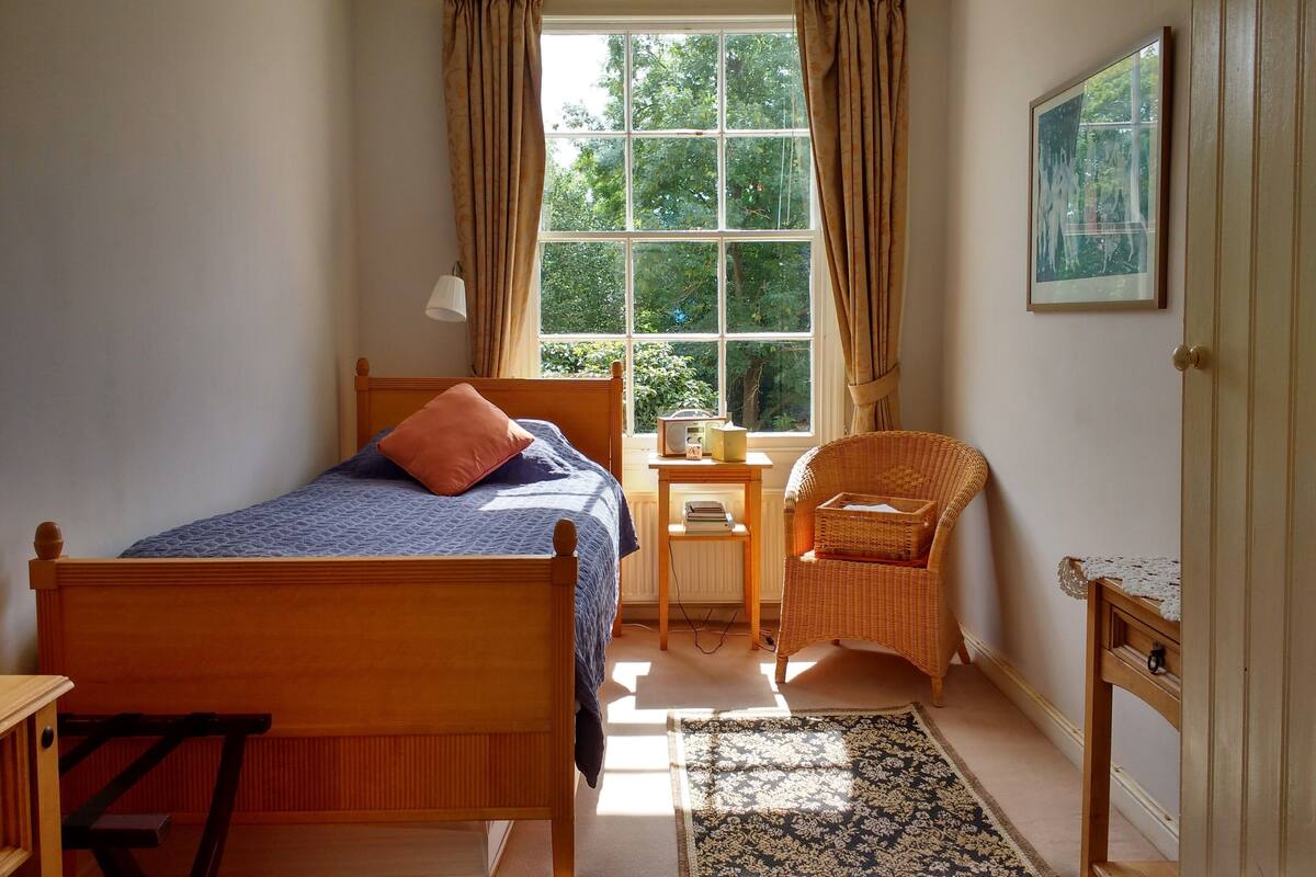 Ein Schlafzimmer mit einem Bett, einem Sessel, einem kleinen Tisch und einem Fenster mit Vorhängen.