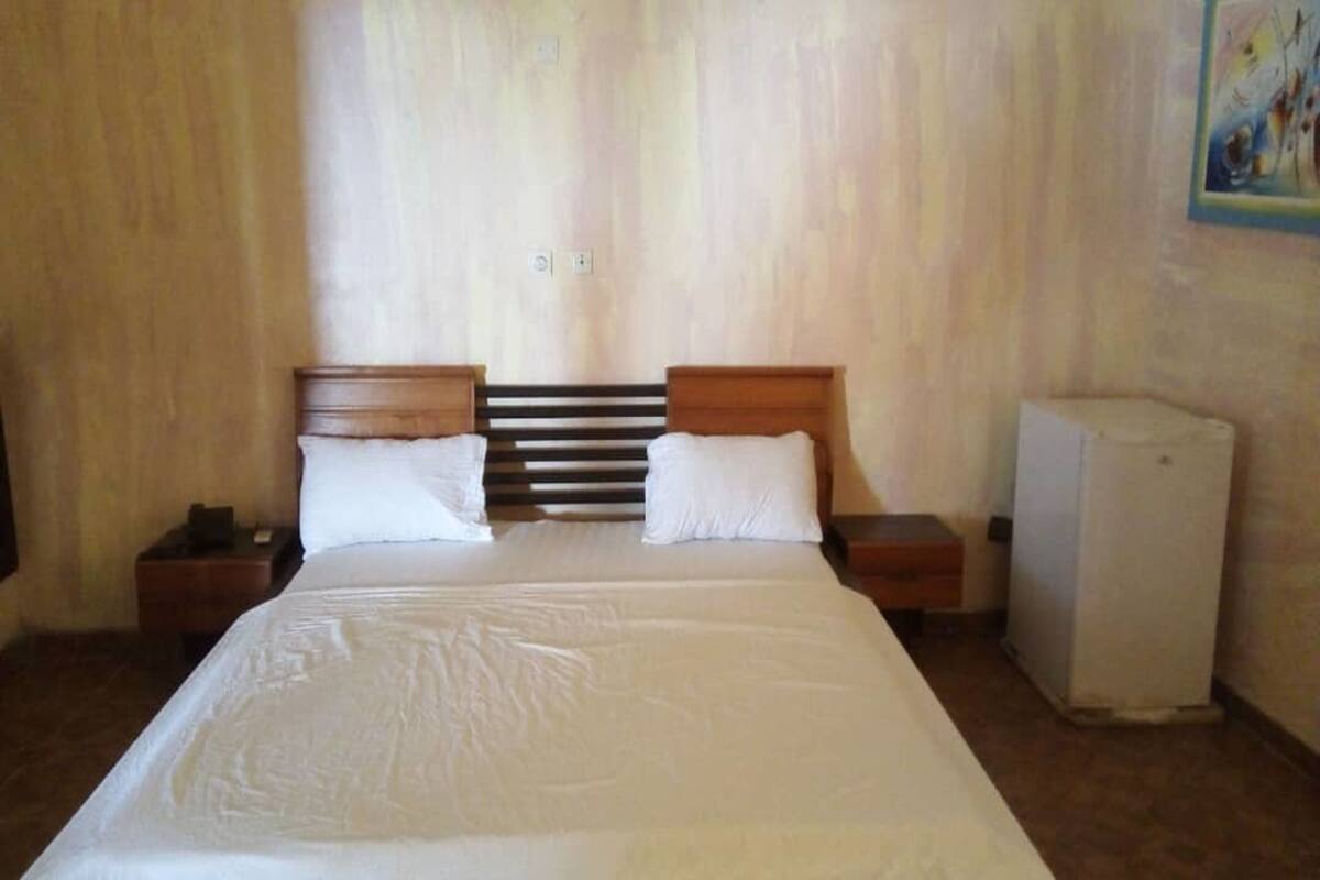 A bedroom with a large bed, two nightstands, a white refrigerator, and a wall-mounted light fixture.