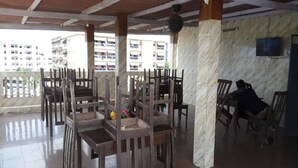A covered outdoor seating area with wooden chairs and tables, a marble wall, and a view of buildings in the background. - Hotel Kama (Abidjan)