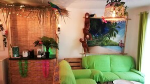 Une pièce avec un canapé vert, une petite table ronde, un mur décoratif orné d’une fresque et une plante suspendue.