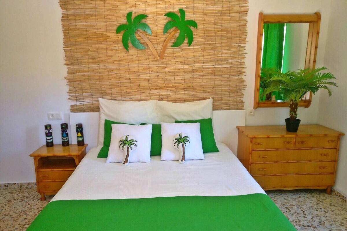 Ein Schlafzimmer mit einem Bett, grünen Kissen, einem Kopfteil mit Palmenmotiven, einem Nachttisch aus Holz, einer Kommode und einem Spiegel.