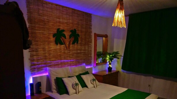 Un lit bien fait, recouvert d’une couvre-lit verte et blanche, un mur à la texture tissée et un luminaire suspendu.