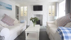 Ein weiß getünchter Raum mit Kamin, zwei weiße Sofas mit Karokissen sowie ein weißer Couchtisch und ein an der Wand befestigter Fernseher.