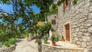 Un sentier de pierres mène à un bâtiment rustique en pierre, doté d’une porte en bois, et est entouré de plantes en pot et de verdure.