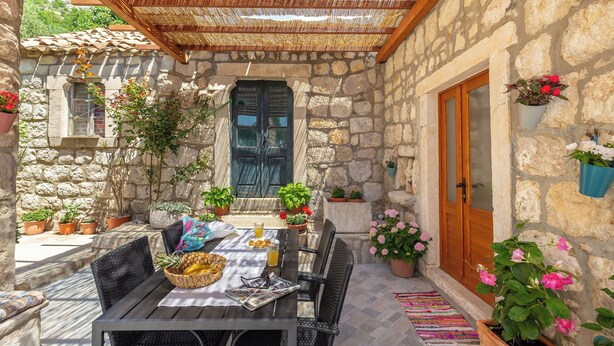 Un espace de restauration en plein air, délimité par des murs de pierre, avec une table dressée pour deux personnes, entourée de plantes en pot et surmonté d’un toit de chaume.