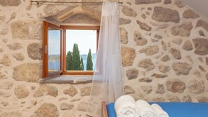 Une pièce aux murs de pierre, avec un lit, une fenêtre donnant sur des arbres et un plan d’eau, et un rideau.