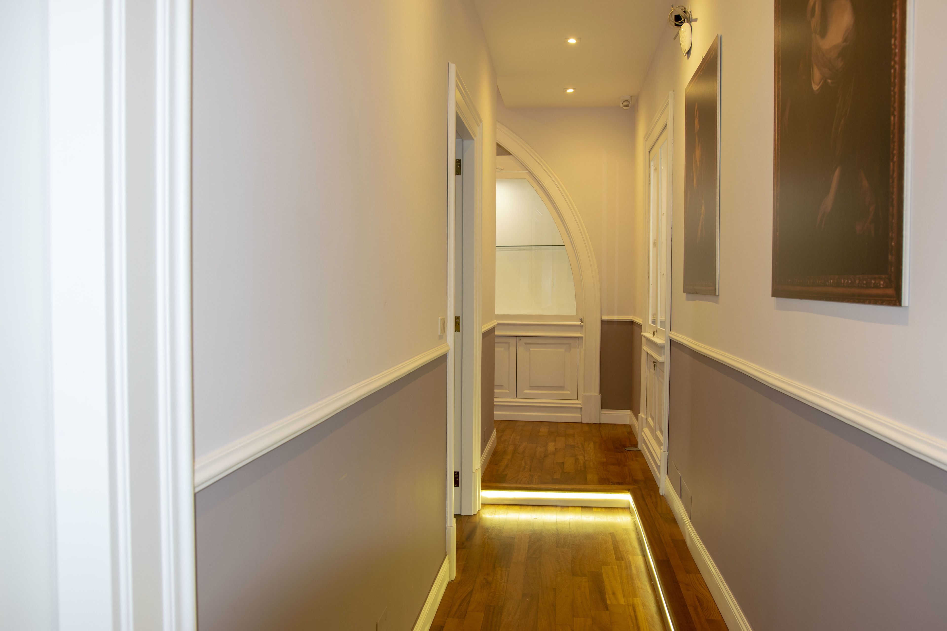 A hallway with a wooden floor, white walls, and a door leading to a room.
