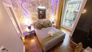A hotel room with a large bed, a nightstand with a lamp, a view of the city through a window, and a decorative wall. - Leone Camere D'Autore (Naples)