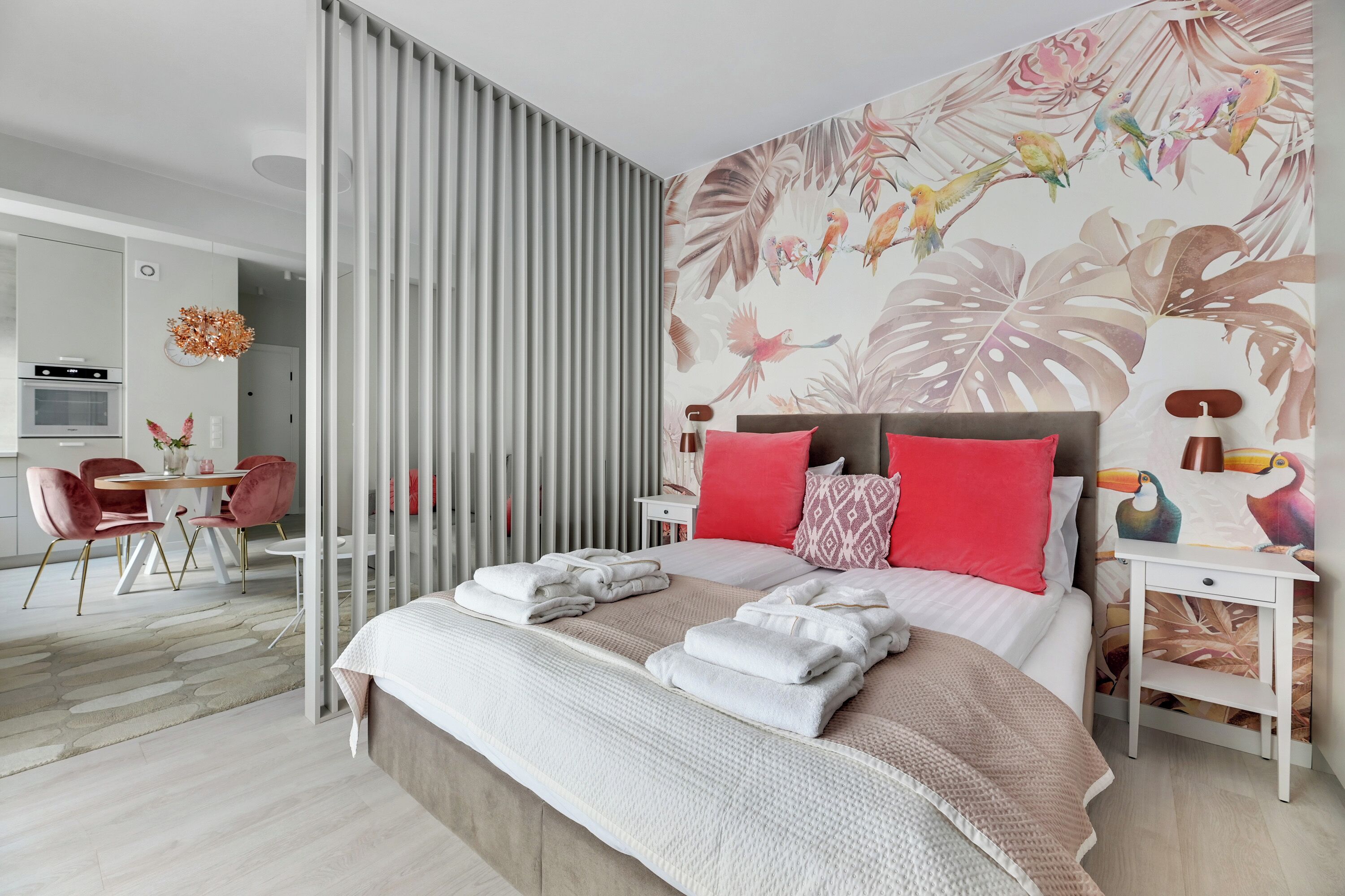 Une chambre moderne avec un grand lit, une table de chevet, un mur recouvert d’un papier peint représentant des oiseaux et des feuilles tropicales, et un coin repas avec une table et des chaises.