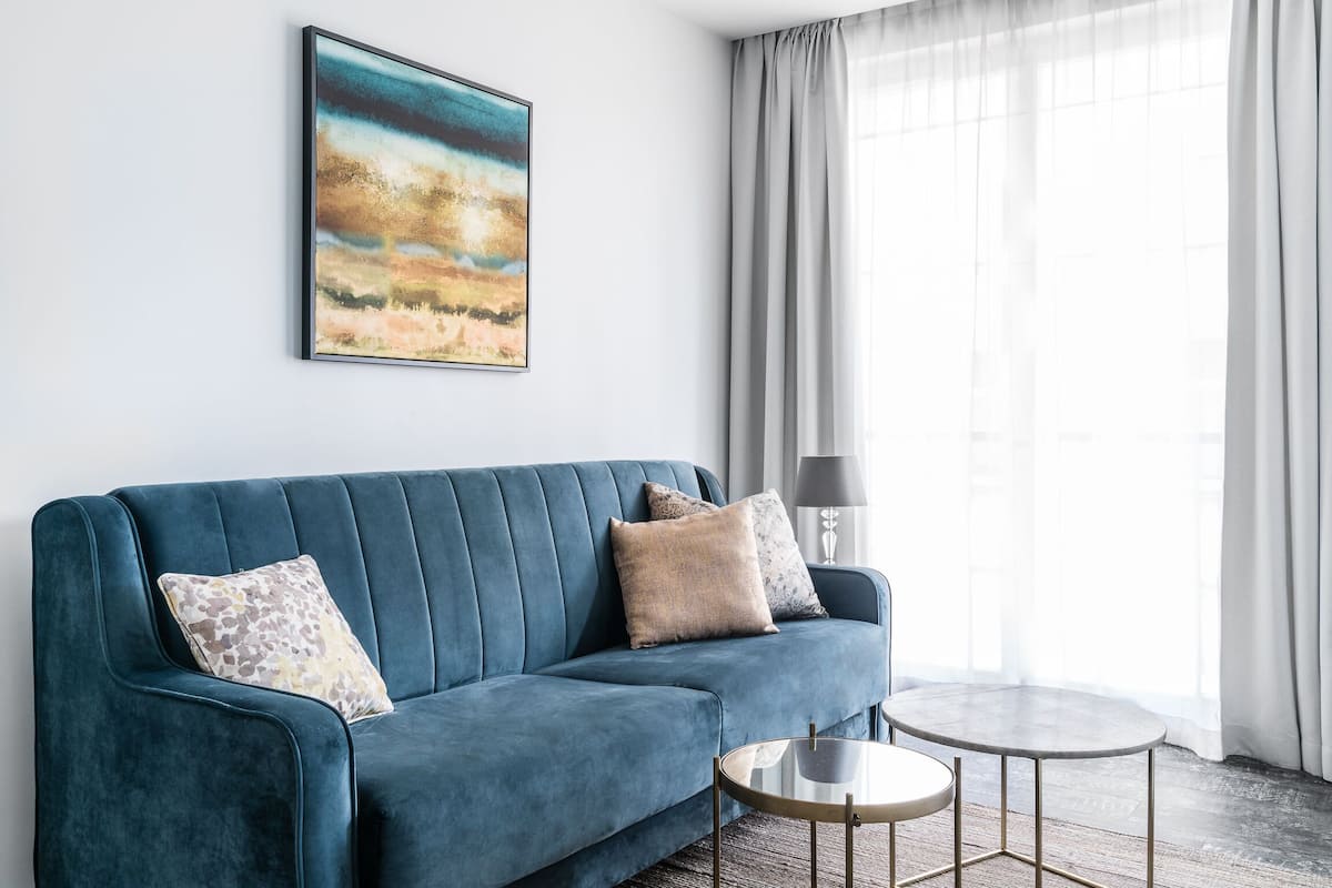 A modern living room with a teal sofa, two patterned cushions, a round coffee table, and a painting on the wall.
