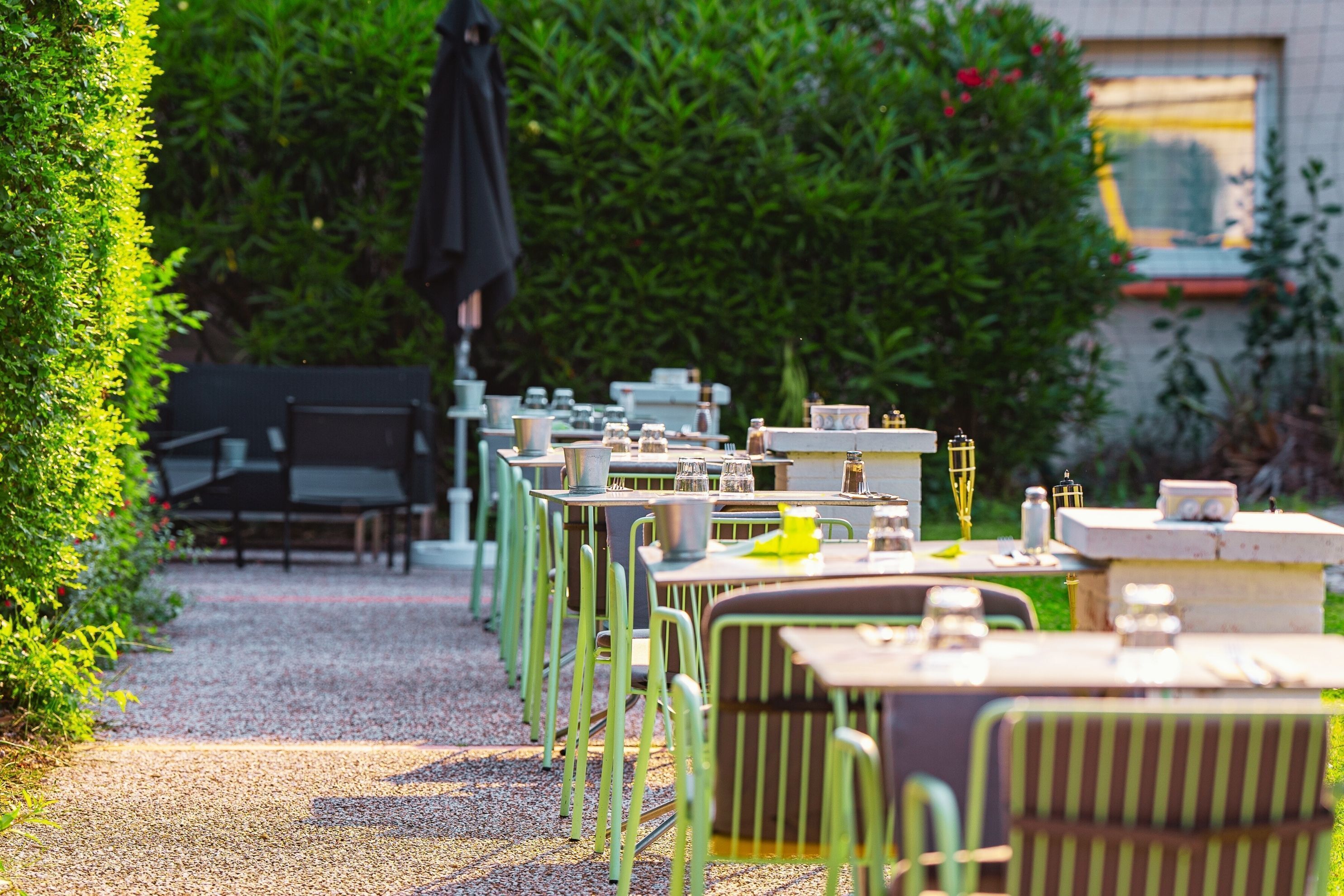 An outdoor dining area with tables and chairs arranged along a gravel path, surrounded by greenery.