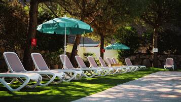 Una fila de sillones blancos con sombrillas verdes, dispuesta sobre una zona cubierta de césped con árboles al fondo.