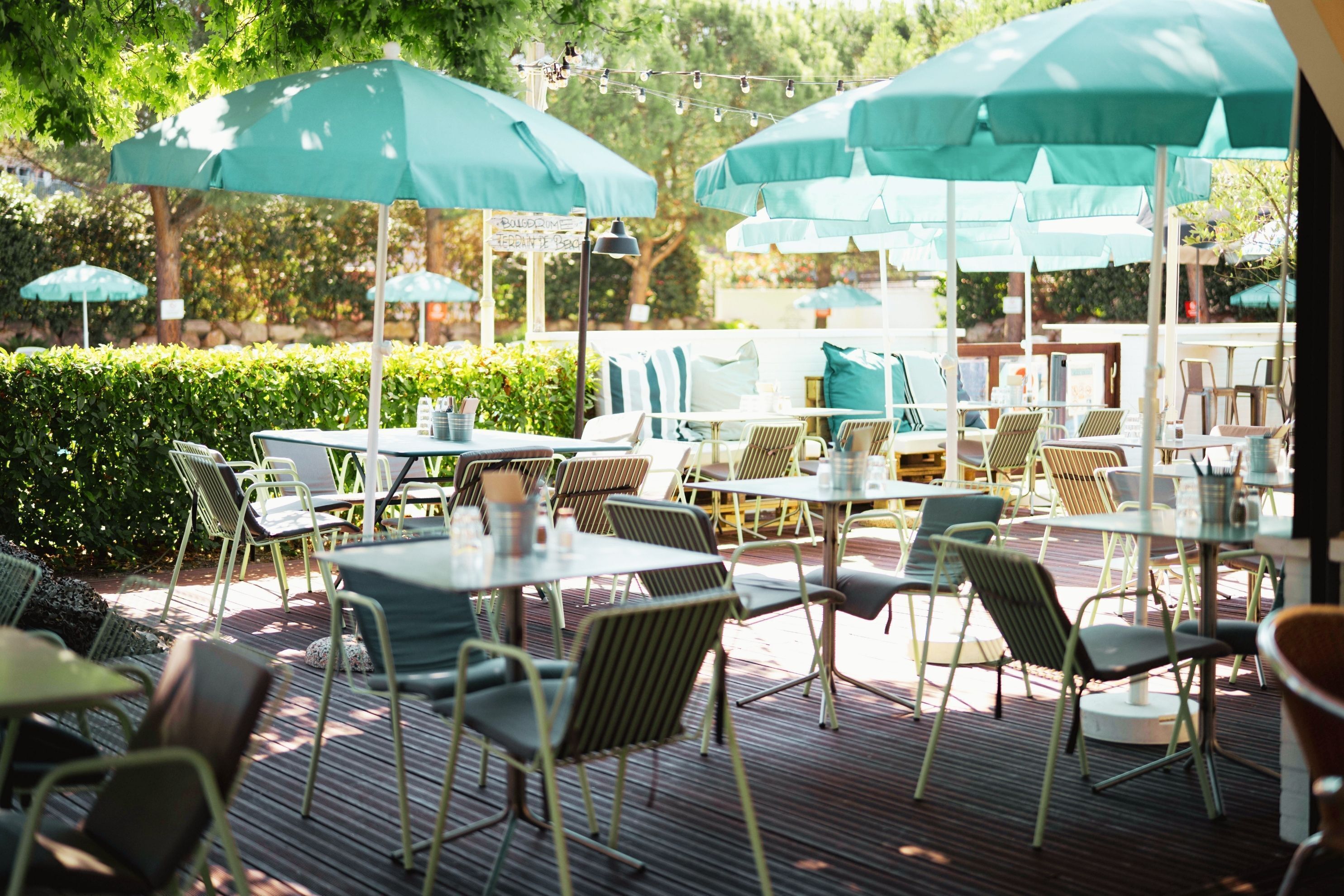 An outdoor patio with tables and chairs, green umbrellas, and a sign indicating a restaurant.