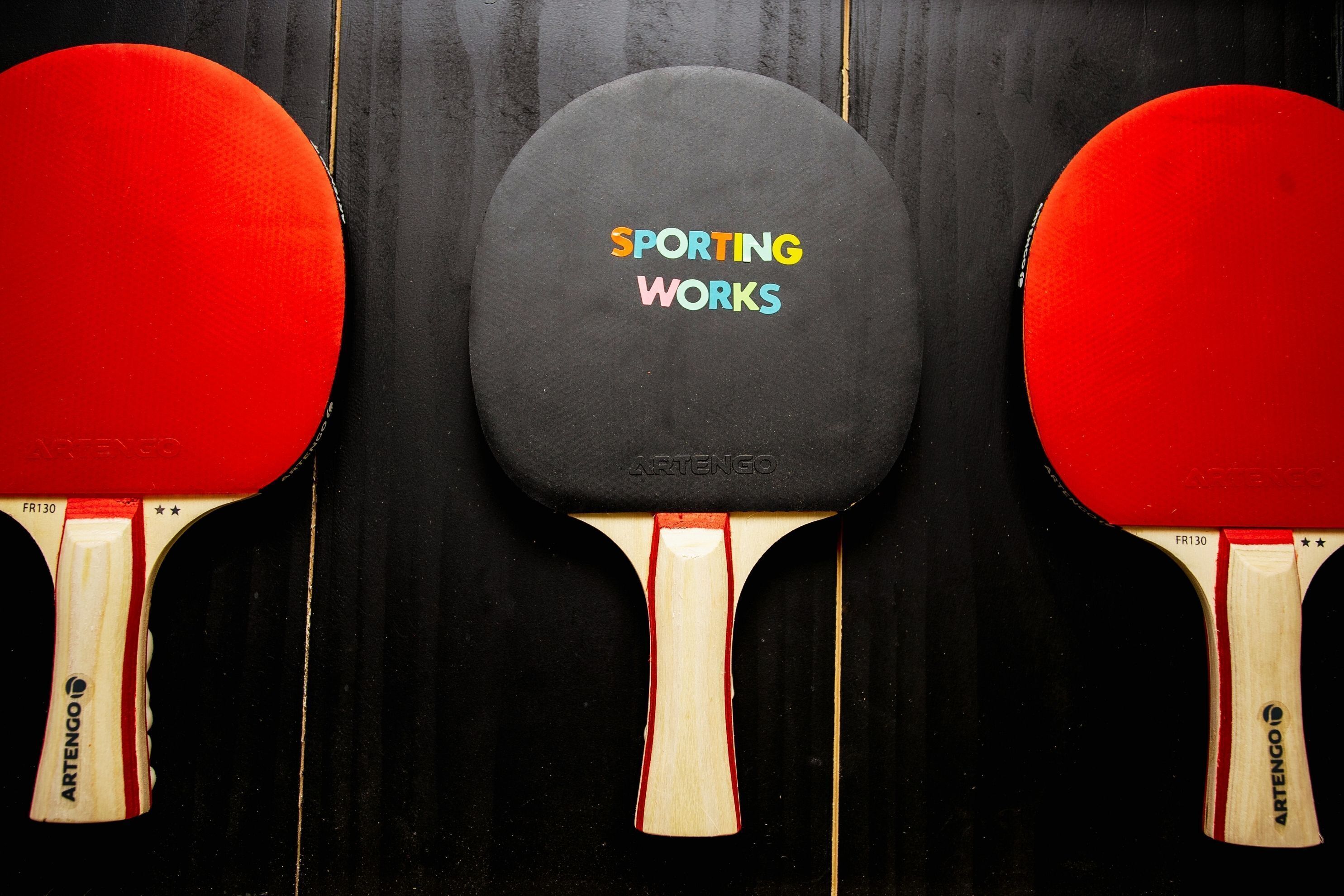 Three table tennis paddles with a black middle paddle labeled "SPORTING WORKS" and two red paddles on either side.