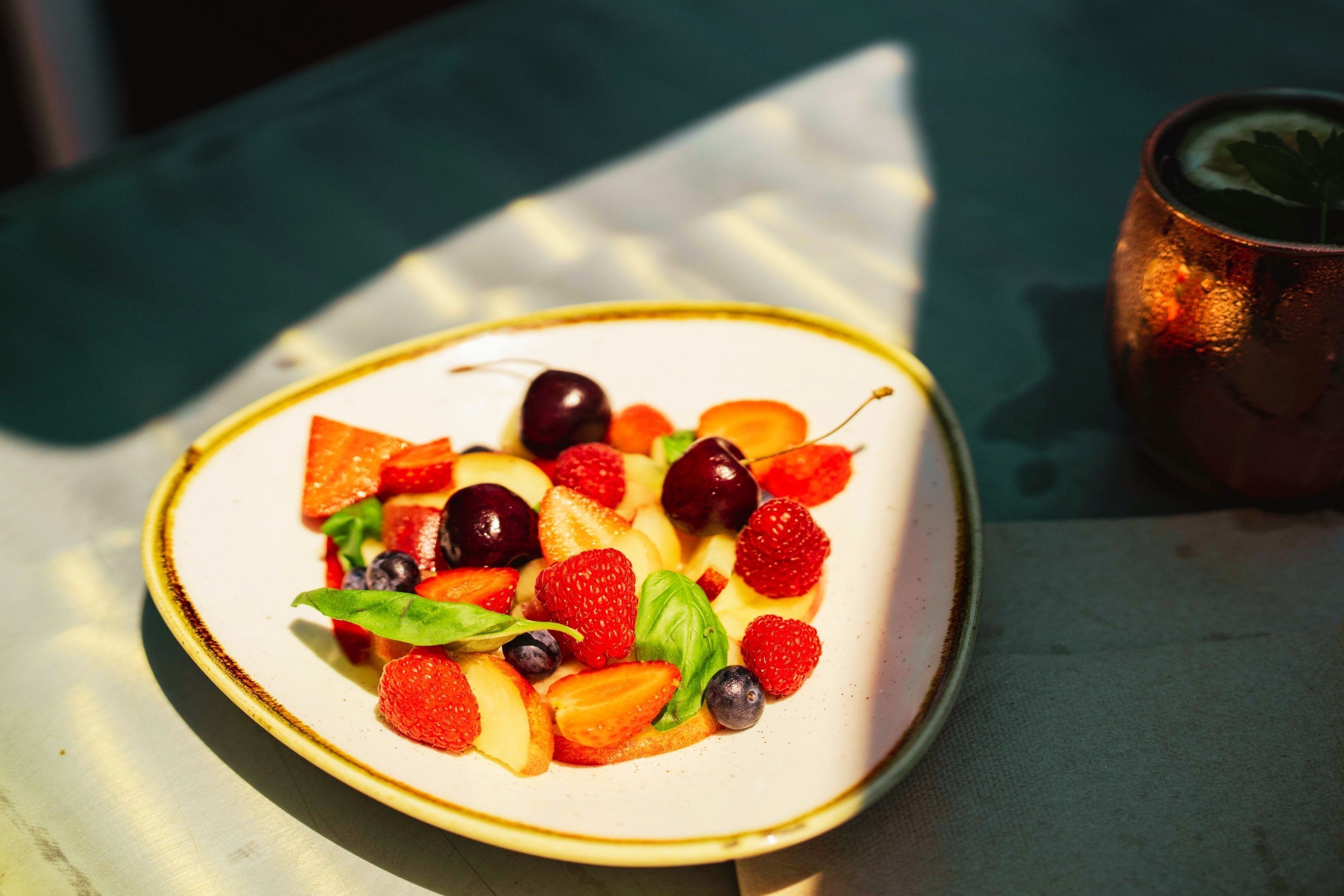 A plate of fruit salad with strawberries, blueberries, and cherries on a white plate with a gold rim, placed on a table with a green cloth.