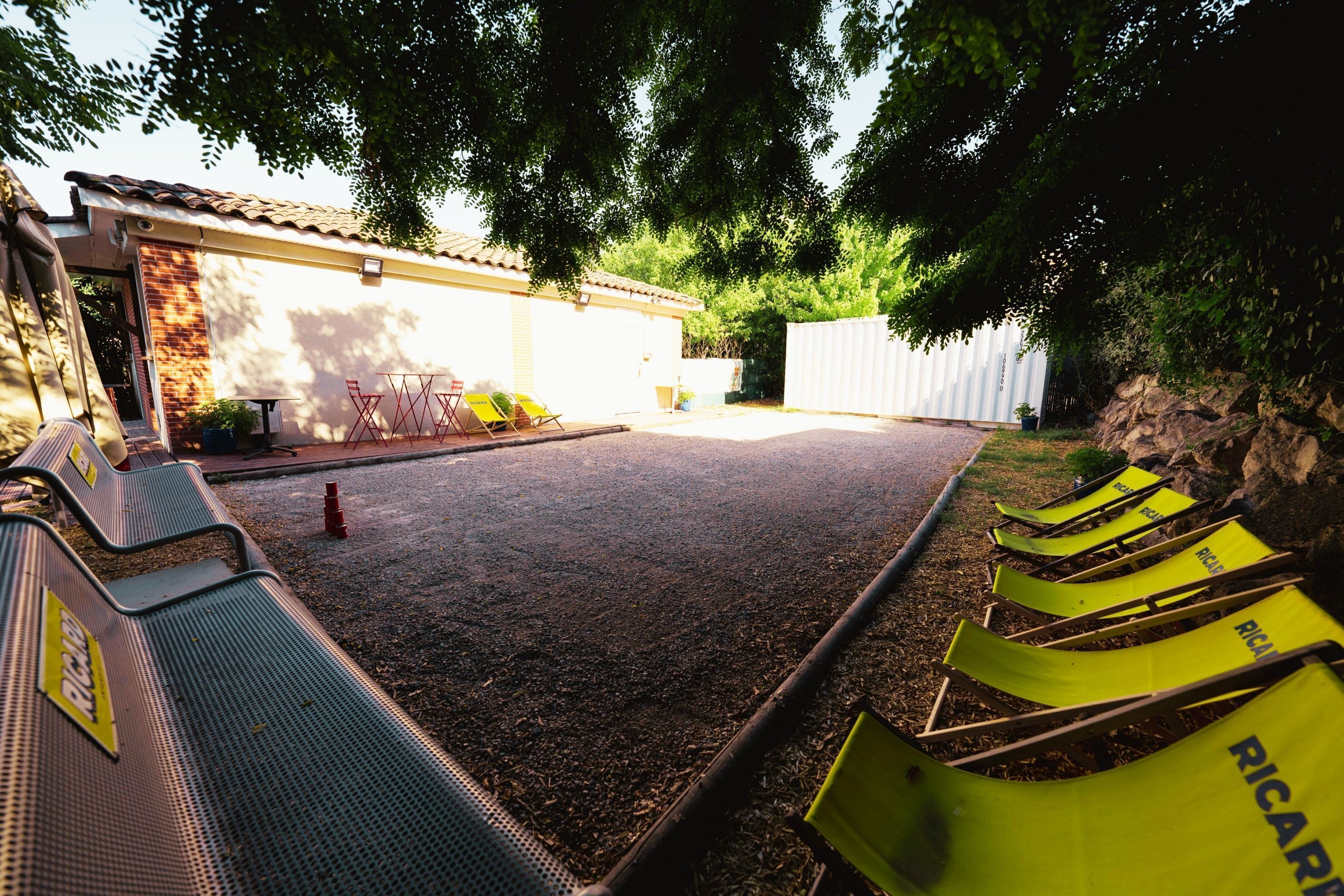 A covered outdoor seating area with metal chairs and tables, a paved floor, and a building with a tiled roof in the background.