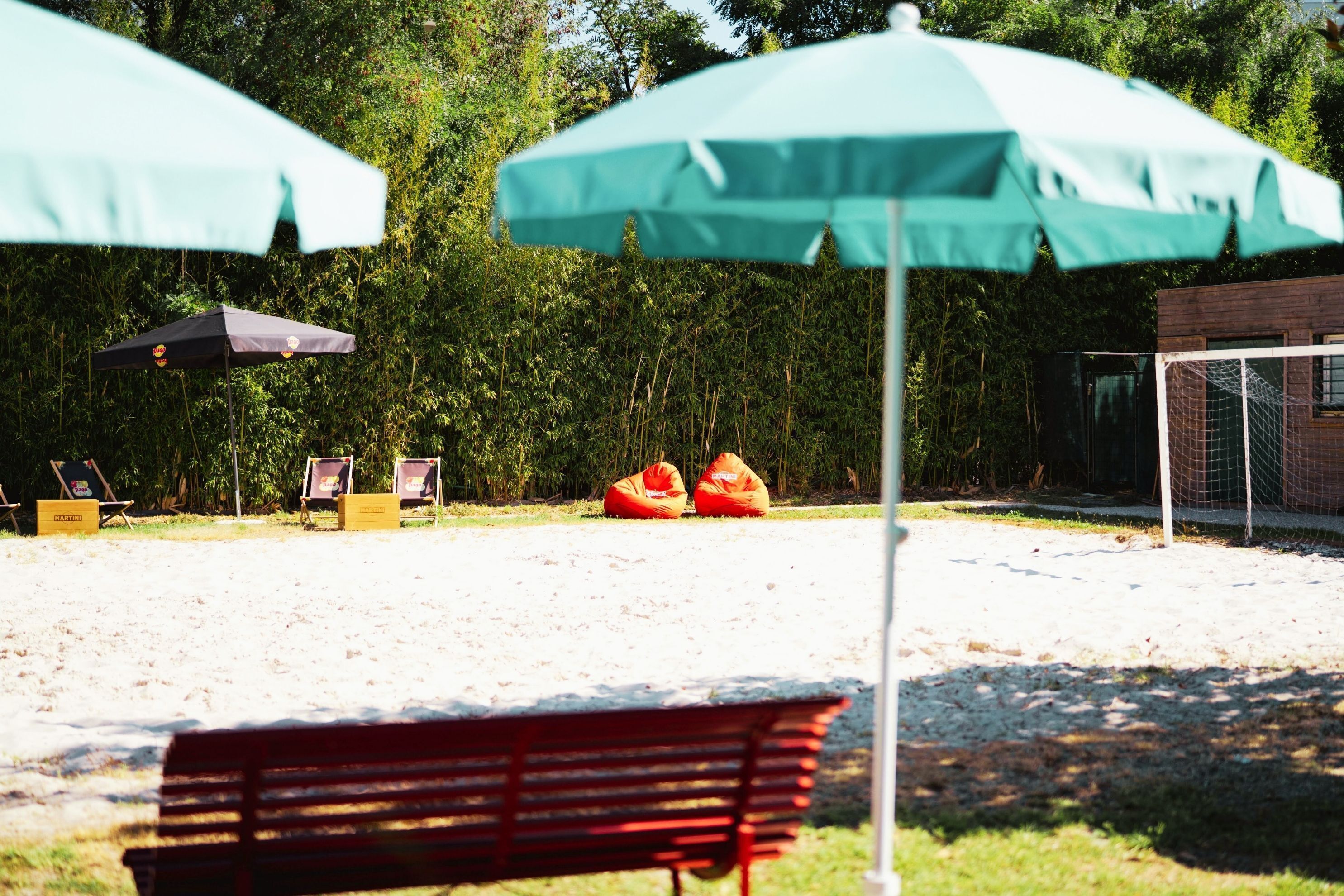A sandy outdoor area with two green umbrellas, a red bench, and two orange bean bags.