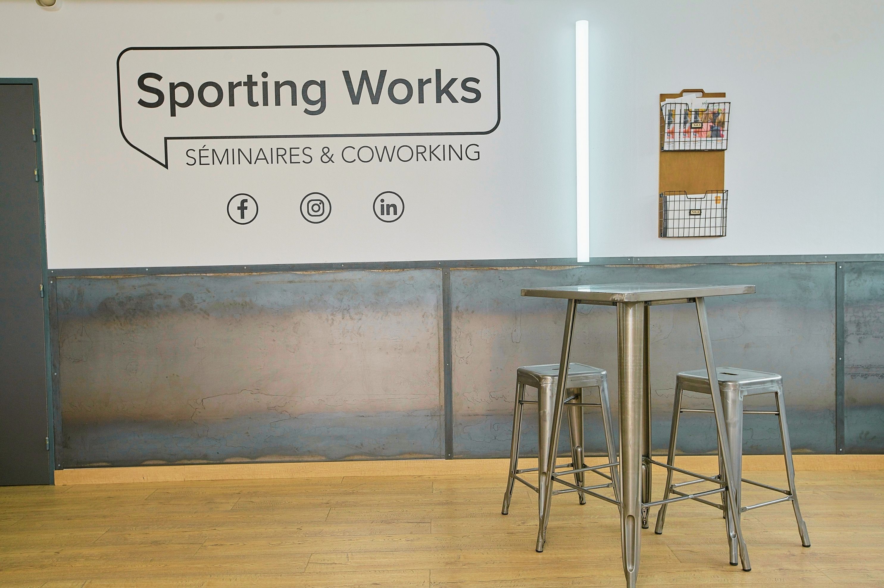 A room with a whiteboard displaying "Sporting Works" and "SÉMINAIRS & COWORKING" along with social media icons. There are two metal stools and a table.