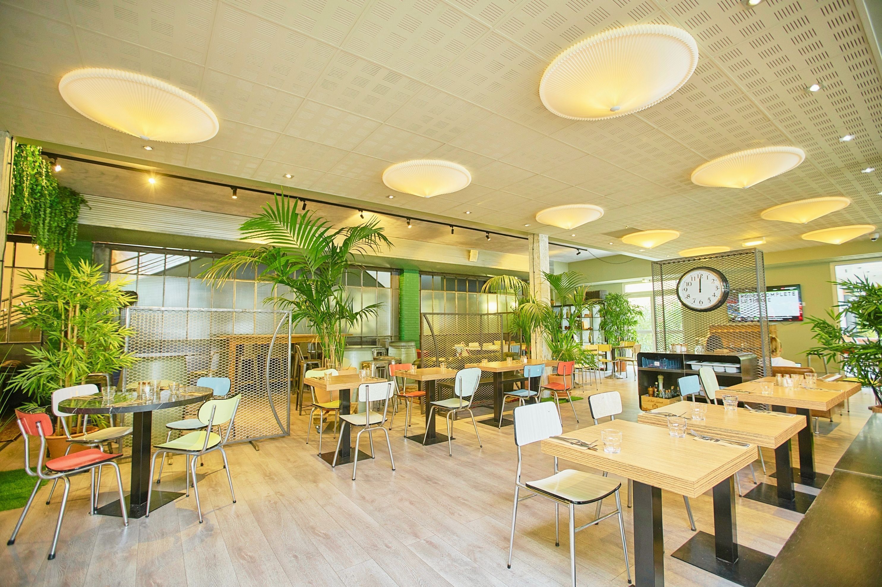 Un vaste espace intérieur aménagé pour les repas, avec de nombreuses tables et chaises, des luminaires au plafond et des plantes en pot.