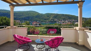 A balcony with two pink chairs, a small table with a glass and fruit, and a view of a lake and mountains. - Hedera Estate, Villa Hedera X (Mokosica)