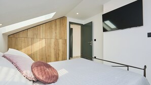 Une chambre moderne avec un plafond en pente, un grand lit doté d’un matelas blanc et d’oreillers roses, une tête de lit en bois, un téléviseur fixé au mur et un puits de lumière.