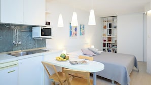 Une cuisine compacte équipée d’un four à micro-ondes, d’un évier et d’un coin repas avec une table et des chaises. On y trouve un lit avec une couvre-lit grise et une bibliothèque contenant divers objets.