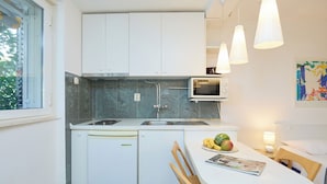 Une cuisine compacte avec des meubles blancs, un four à micro-ondes encastré et un lave-vaisselle. On y trouve une table à manger avec des chaises, un panier de fruits et un magazine.