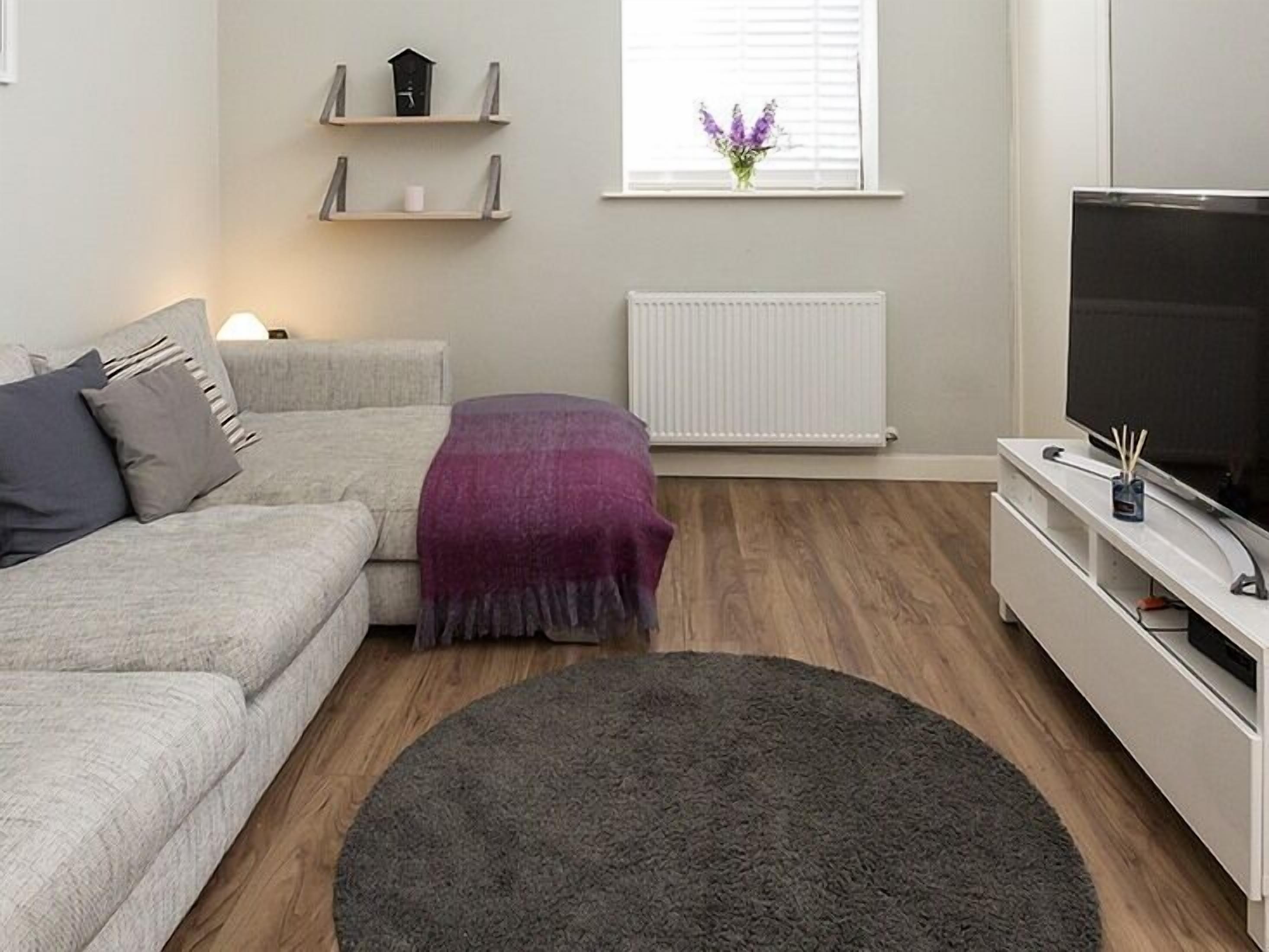 A modern living room with a grey sofa, a dark grey rug, a white TV stand, and a window with a view of purple flowers.