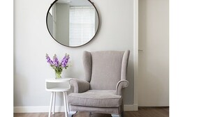 A grey armchair with a round mirror above it, a small white side table with a vase of purple flowers, and a wooden floor. - The Lane Apartment (Edinburgh)