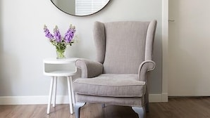 A room with a grey armchair, a small white side table with a vase of purple flowers, and a round mirror on the wall. - The Lane Apartment (Edinburgh)