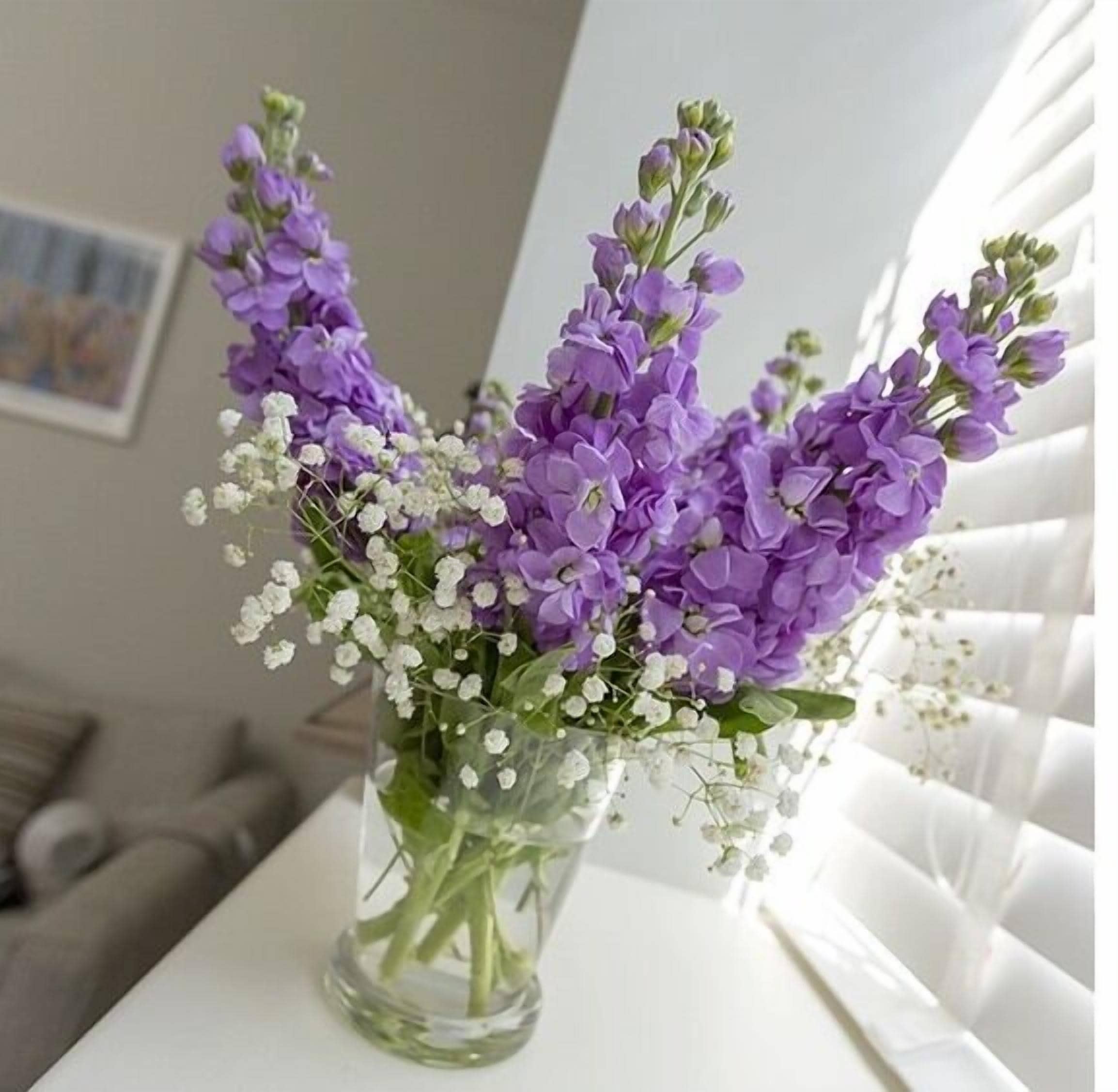 A clear glass vase with purple flowers and small white flowers on a white surface.
