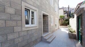 A narrow, paved courtyard with stone walls and a green door, leading to a building with multiple windows and steps.