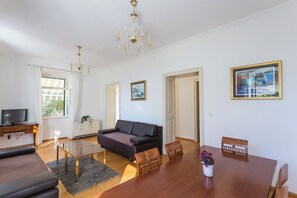 A modern living room with a sofa, a glass coffee table, a dining table with chairs, a TV on a stand, and framed pictures on the walls.