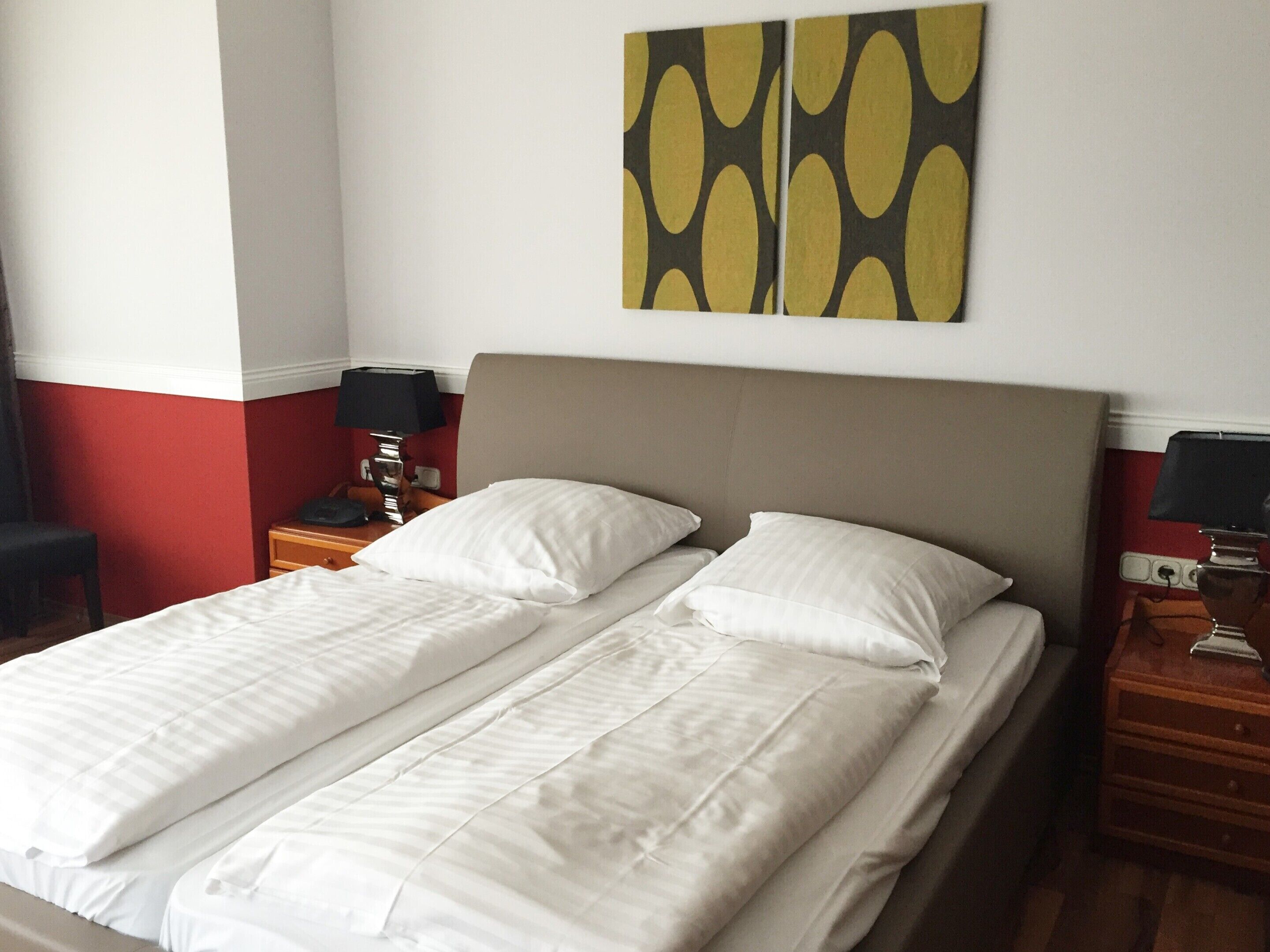 A double bed with white linens, a wooden nightstand with a lamp, and a wall with a geometric painting.