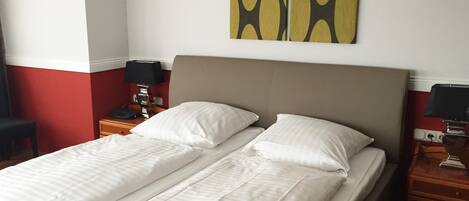 A double bed with white linens, a wooden nightstand with a lamp, and a wall with a geometric painting.