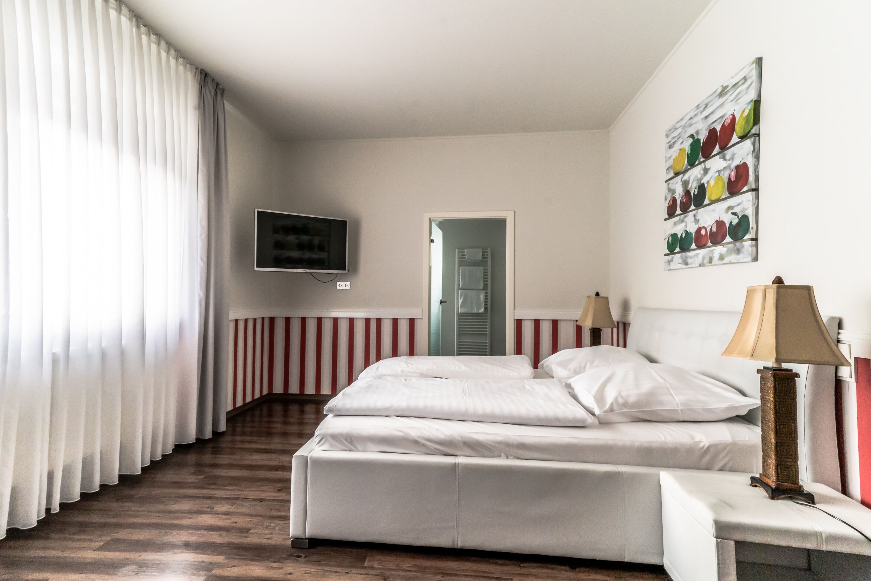 A hotel room with a bed, a TV mounted on the wall, a door leading to another room, and a painting of fruits on the wall.