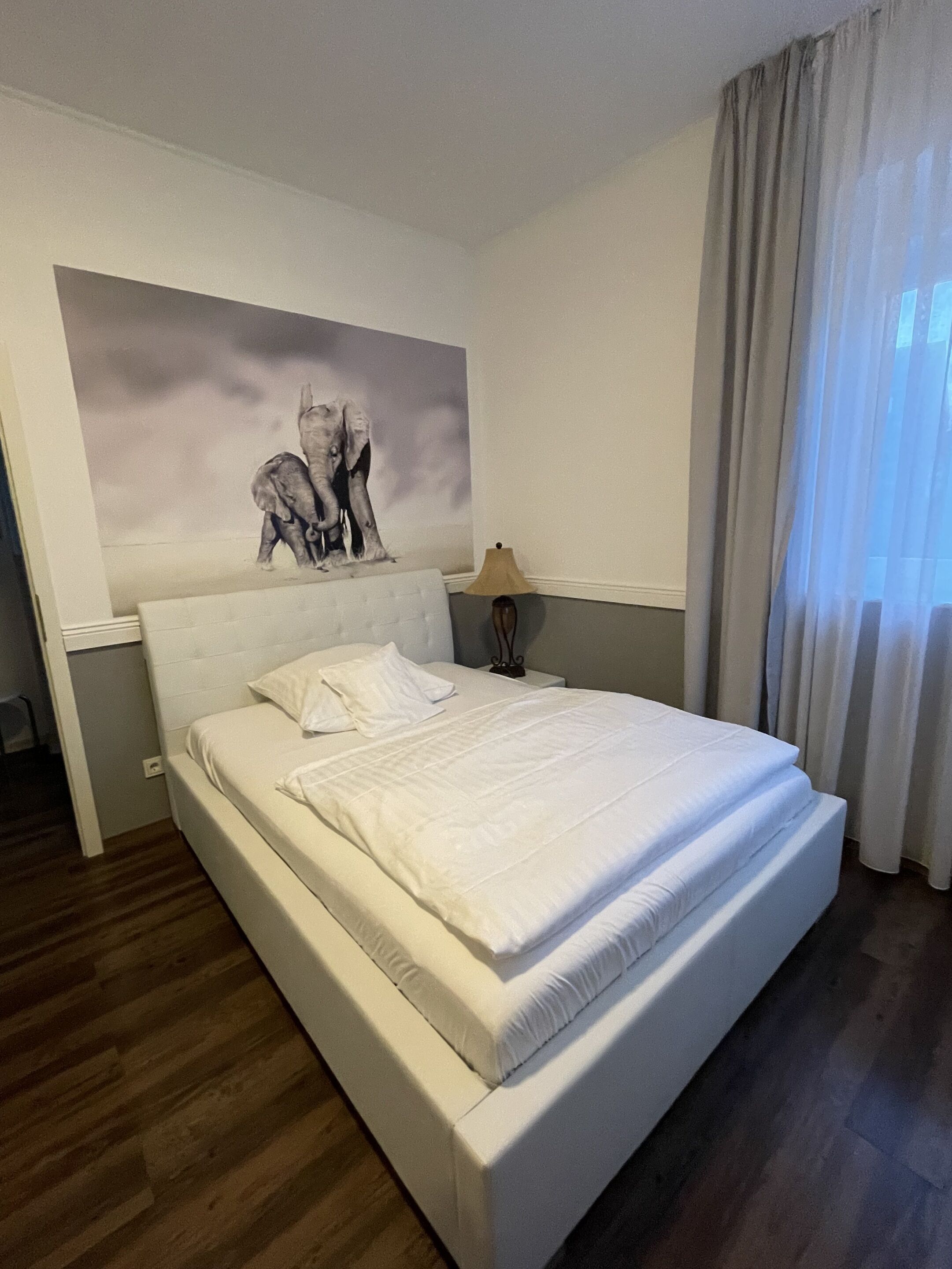 Una cama bien hecha en una habitación de hotel con un cuadro grande en la pared, una lámpara y una ventana con cortinas.