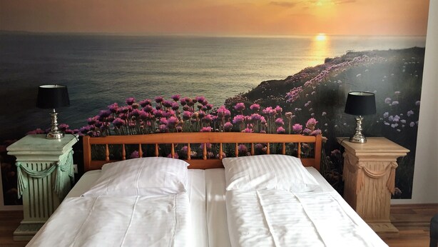 Een slaapkamer met een bed, twee nachtkastjes en een muurschildering van een zonsondergang boven de oceaan.
