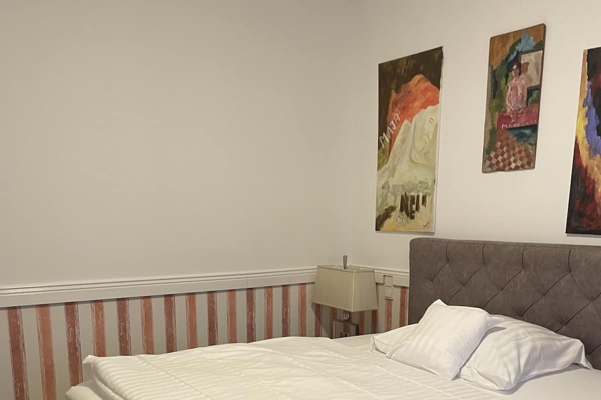 Una cama bien tendida con un cabecero gris, una pared a rayas y una lámpara sobre una mesita de noche.