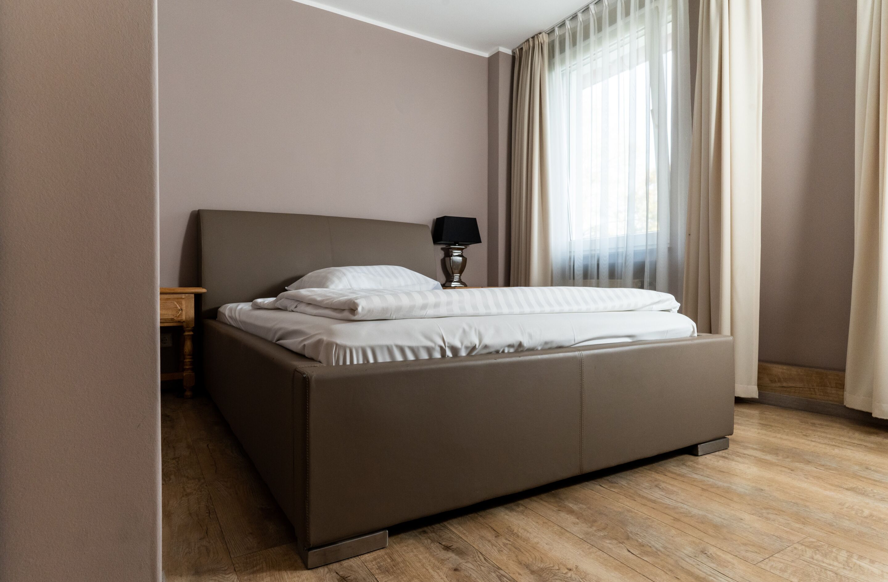 A modern hotel room with a large bed, a bedside table with a lamp, and a view through sheer curtains.