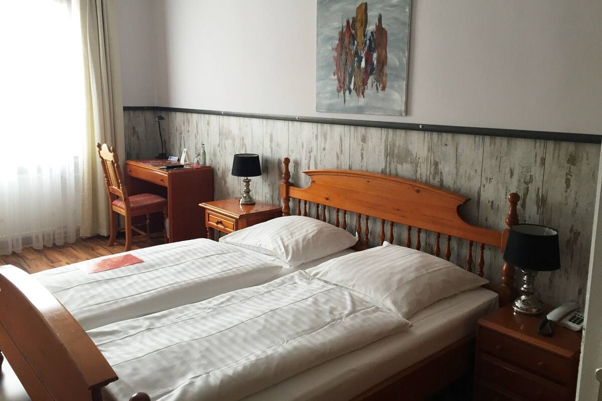 Habitación de hotel con una cama de madera, un escritorio con silla, una lámpara y un cuadro en la pared.