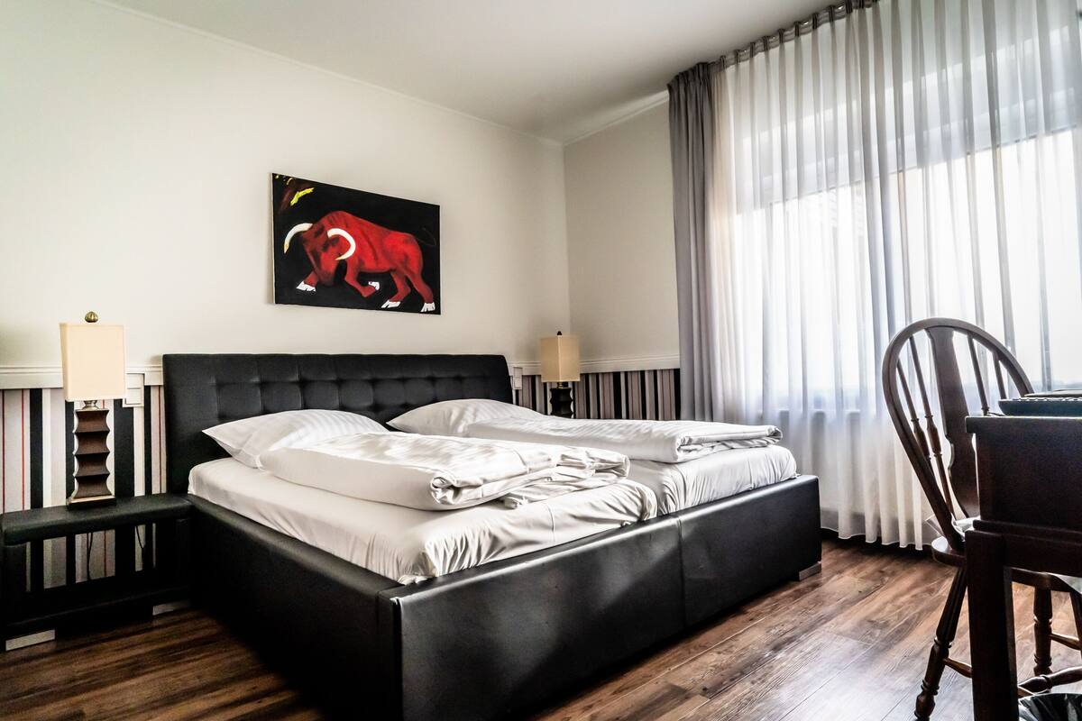 Habitación de hotel con una cama tapizada de negro, ropa de cama blanca, un cuadro de un elefante, suelo de madera, un escritorio y una silla.