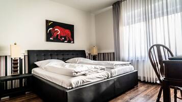 A hotel room with a black upholstered bed, white linens, a painting of an elephant, a wooden floor, a desk, and a chair.
